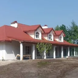 Nowy, czerwony dach z dachówki ceramicznej na parterowym domu z białymi ścianami i oknami w lukarnach. W tle drzewa.