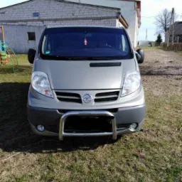 Szary van Nissan z chromowanym orurowaniem przedniego zderzaka, stojący na trawie przed budynkiem gospodarczym w słoneczny dzień.