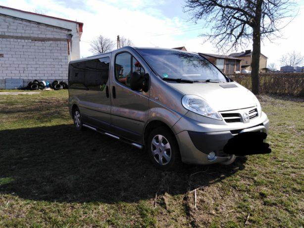 Szary van Nissan Primastar na trawiastym terenie, z budynkiem w tle i stosem opon.