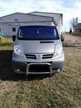 Szary van Nissan z chromowanym orurowaniem przedniego zderzaka, stojący na trawie przed budynkiem gospodarczym w słoneczny dzień.