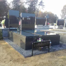 Czarny, polerowany pomnik z krzyżem na cmentarzu, otoczony kostką brukową i niską metalową ławeczką, w tle drzewa i inne nagrobki.