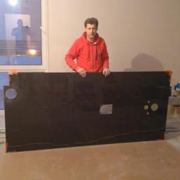 Usługi remontowo-budowlane Jakub Jurewicz - Mężczyzna w czerwonej bluzie trzyma czarny blat z otworami na umywalkę i armaturę, na podłodze narzędzia i materiały budowlane, w tle okno z odbiciem osoby robiącej zdjęcie.