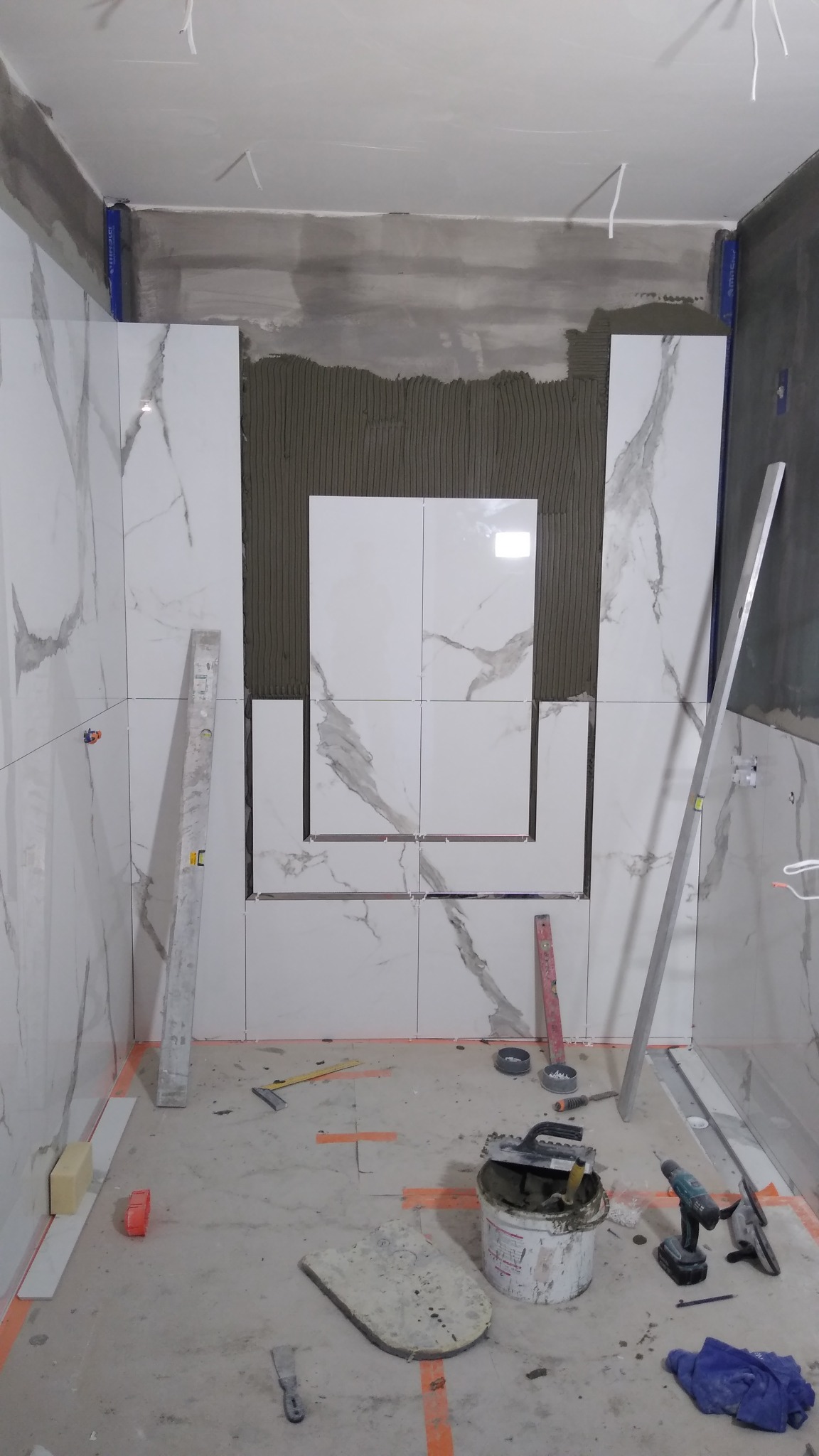 Montaż białych płytek imitujących marmur na ścianie w łazience, widoczne narzędzia, poziomica i klej do płytek, etap prac wykończeniowych.