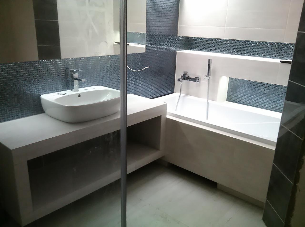 Nowoczesna łazienka z białą umywalką na blacie z półkami, mozaiką na ścianie i wanną obudowaną jasnymi płytami.