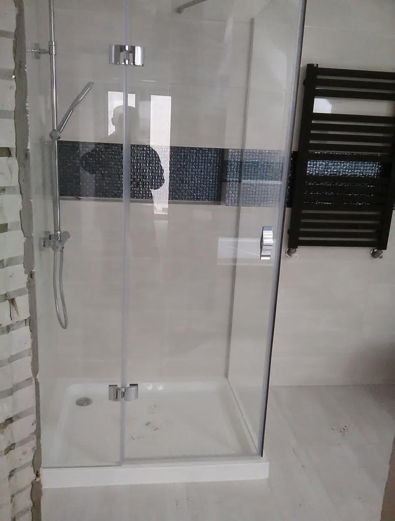 Nowoczesna kabina prysznicowa z chromowanymi detalami, widoczna mozaika dekoracyjna na ścianie i czarny grzejnik drabinkowy, fragment nieotynkowanej ściany po lewej stronie.