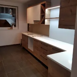 Nowoczesna kuchnia z drewnianymi frontami szafek i białym blatem, oświetlenie LED pod szafkami wiszącymi, widok na okno z widocznym wnętrzem innego pomieszczenia.