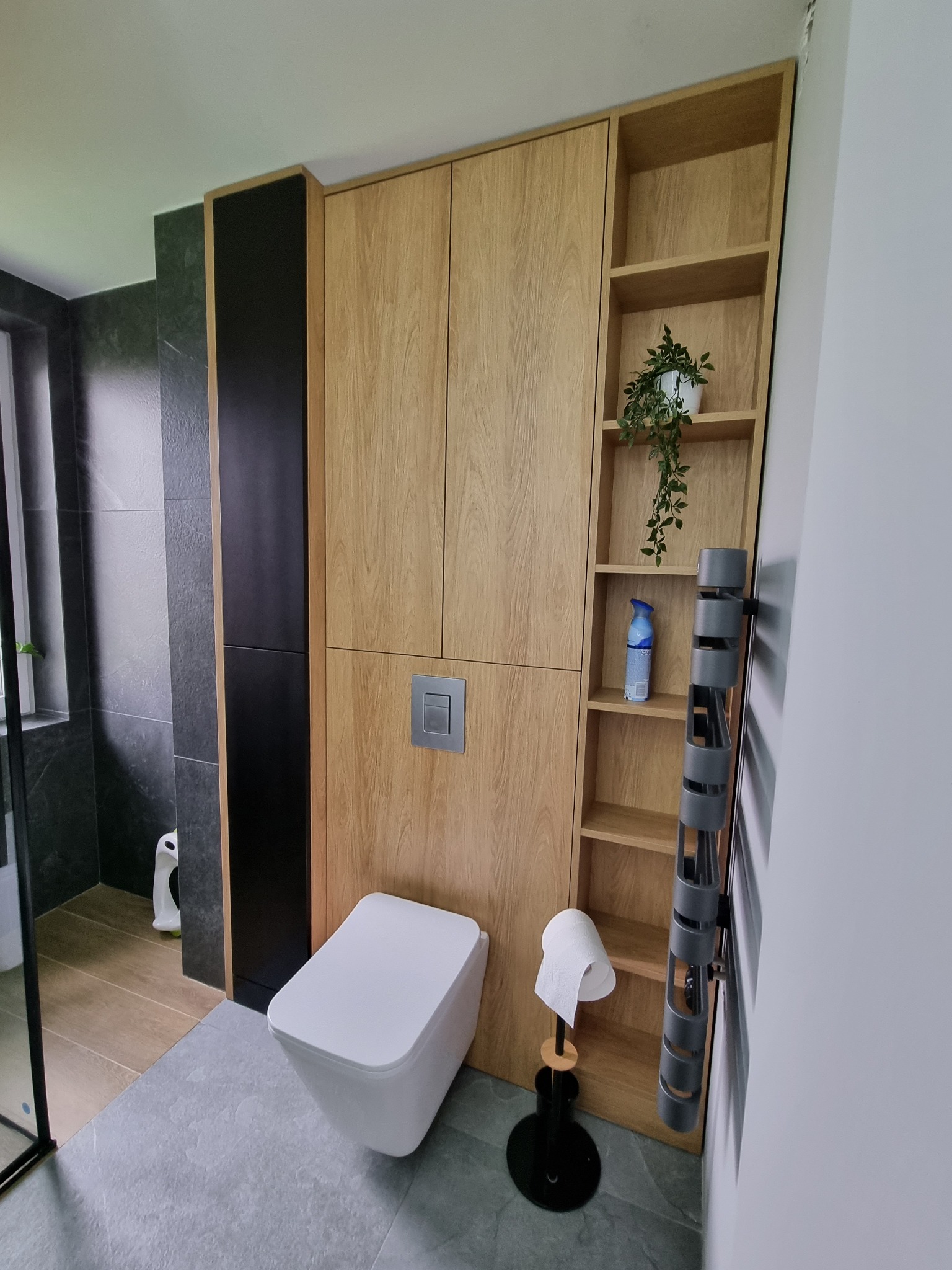 Nowoczesna toaleta podwieszana z zabudową z drewnopodobnej płyty, zintegrowana z czarnym panelem i otwartymi półkami, na których stoi roślina i odświeżacz powietrza. Szary grzejnik drabinkowy obok.