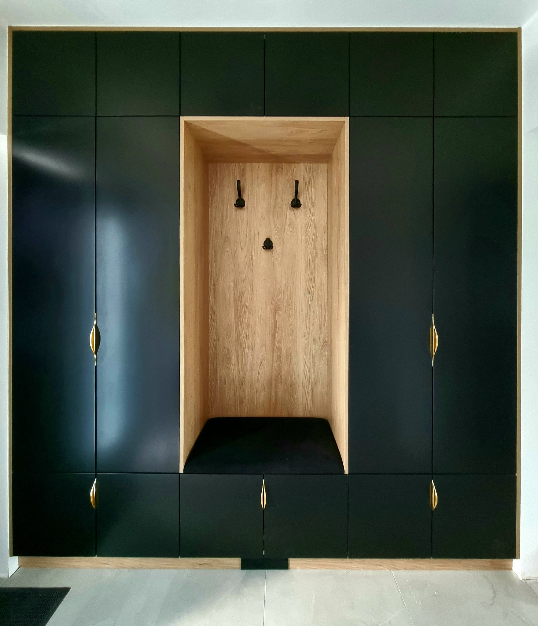 Czarna zabudowa meblowa z drewnianą wnęką z siedziskiem i wieszakami, uchwyty w kolorze złotym.