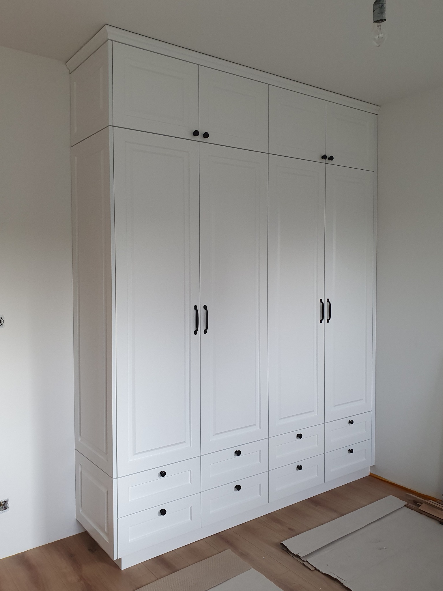 Wysoka, biała szafa z górnymi szafkami i dolnymi szufladami, czarne okucia, widok z dołu, podłoga częściowo zakryta kartonem zabezpieczającym.