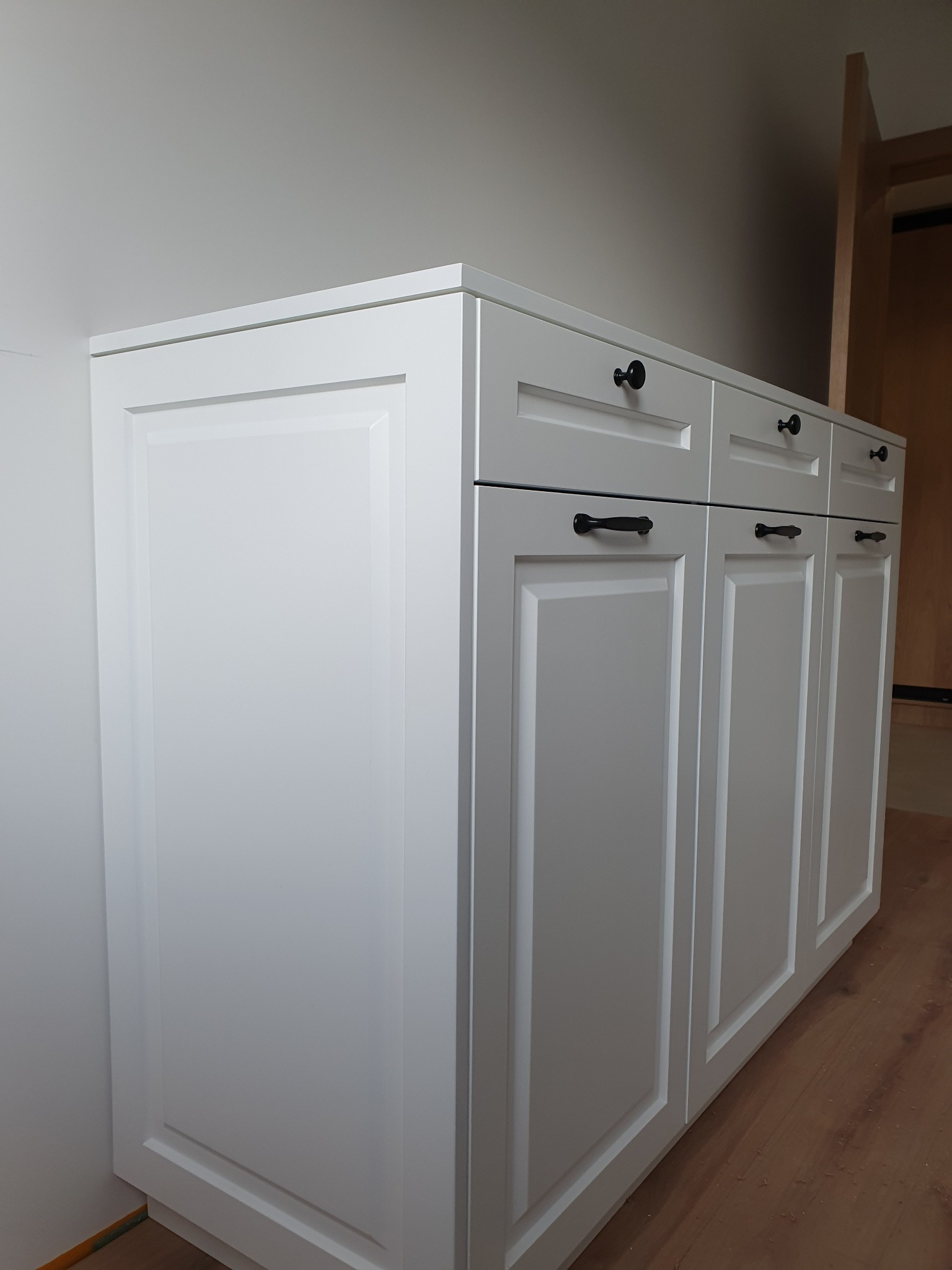 Biała szafka kuchenna z frezowanymi frontami i czarnymi uchwytami, widok z boku na tle drewnianej podłogi.