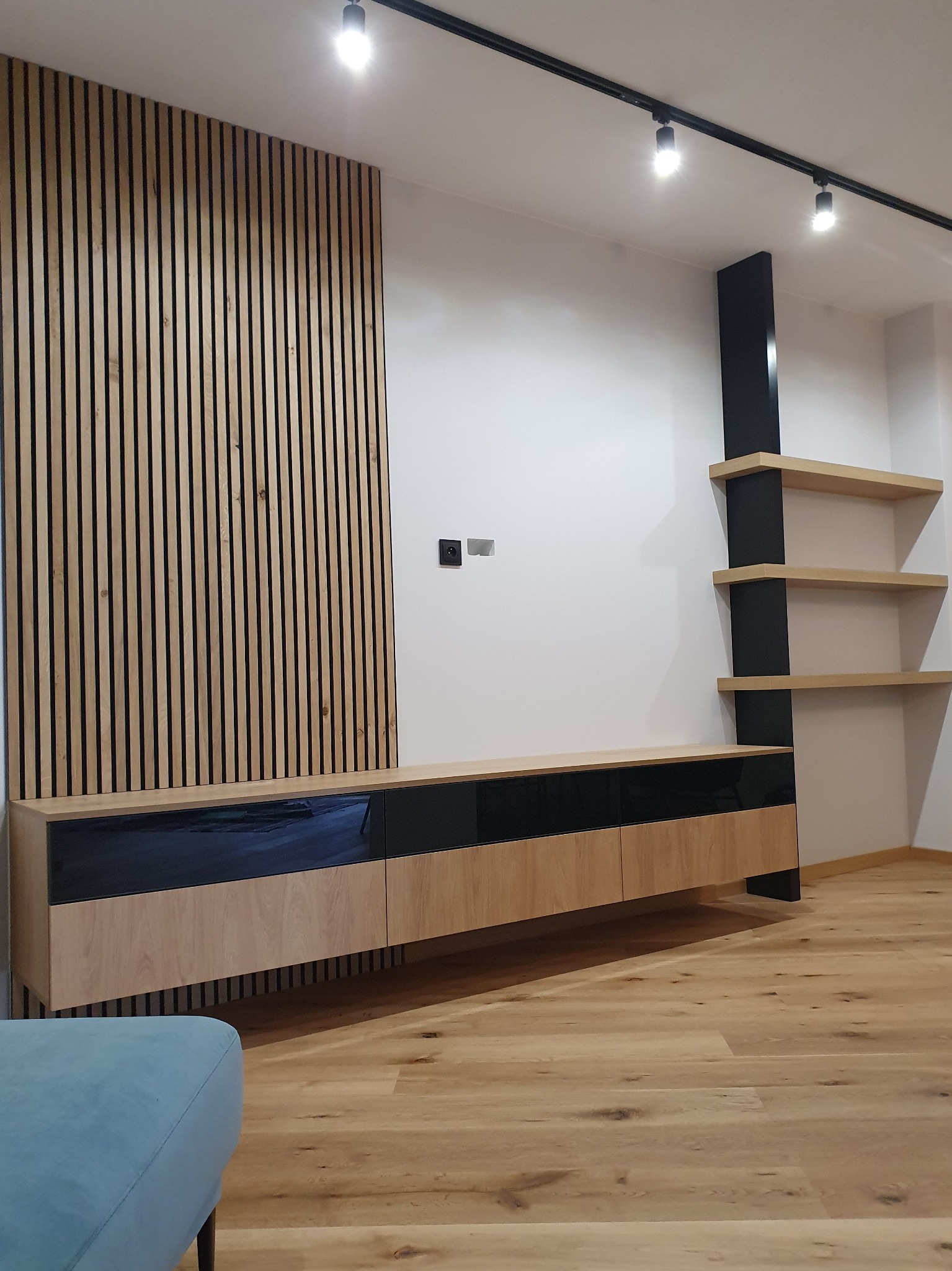 Nowoczesny salon z drewnianą ścianą z lamelami, podwieszaną szafką RTV z czarnymi frontami na wysoki połysk, oraz półkami na czarnym wsporniku.