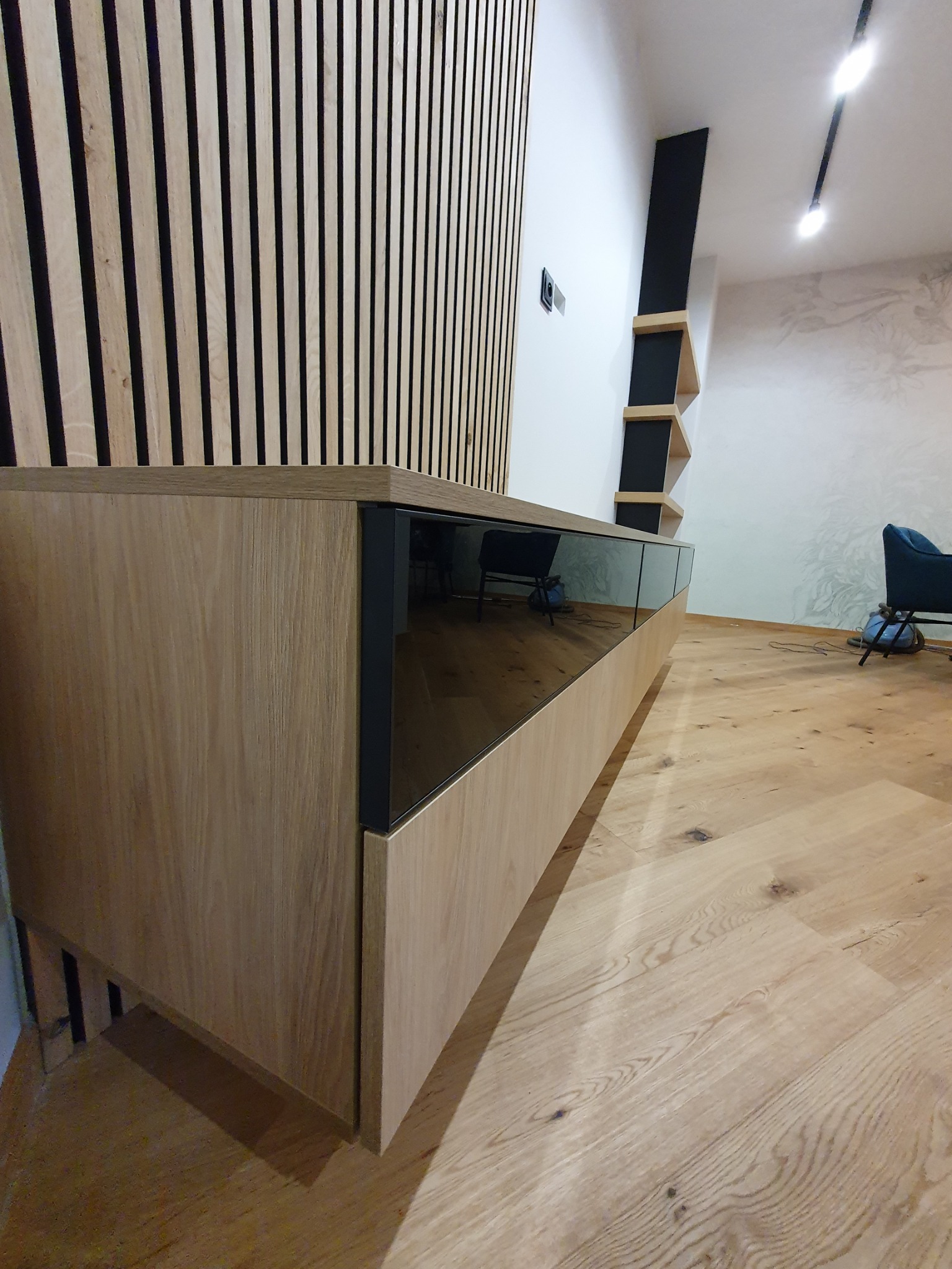 Nowoczesna szafka RTV na wymiar z drewnianym fornirem i czarnymi szklanymi frontami, dopasowana do drewnianej ściany z lameli i podłogi, z czarnymi półkami na ścianie w tle.