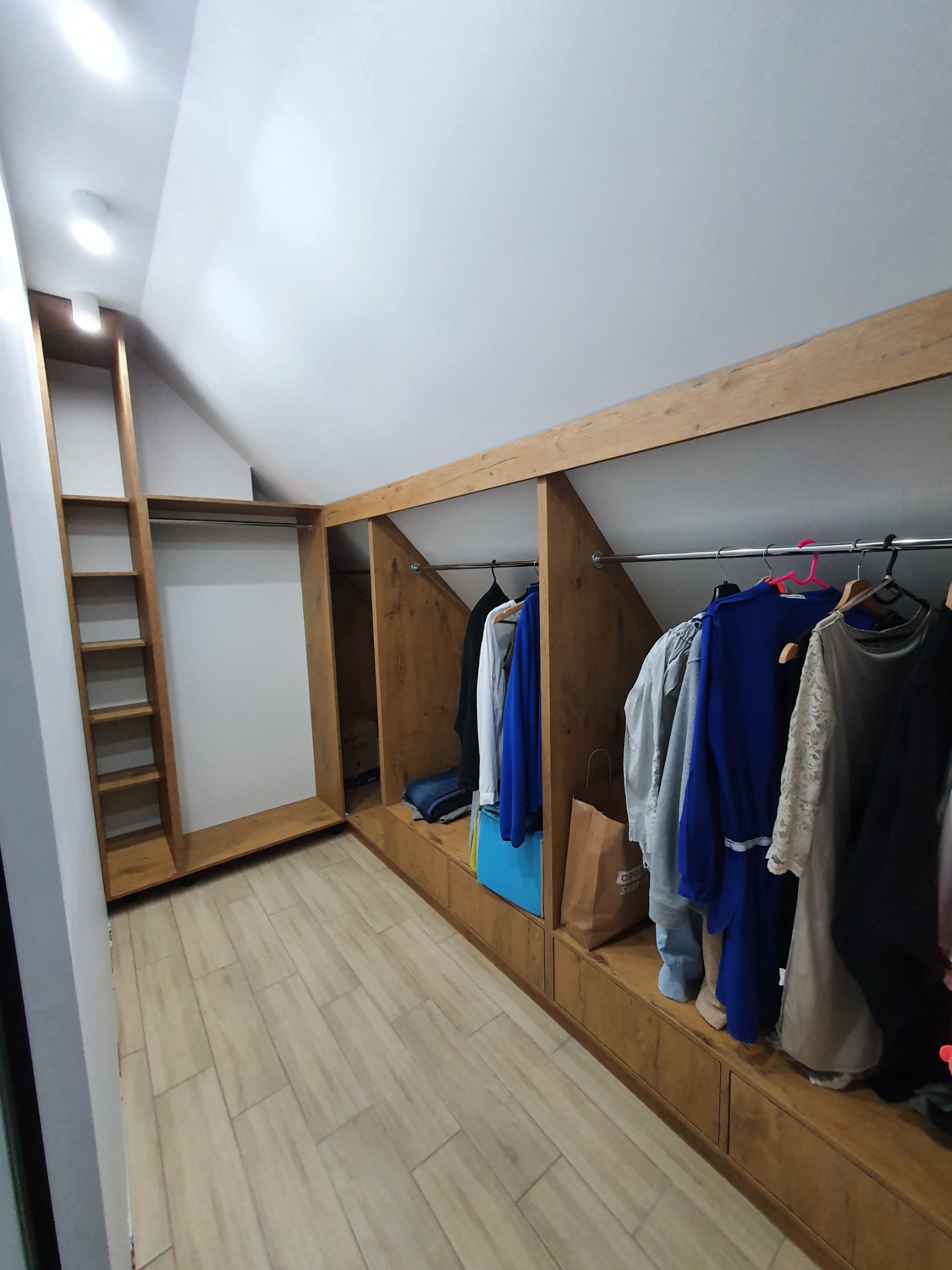 Drewniana garderoba w skosie na poddaszu, z półkami i wnękami na ubrania, podłoga wyłożona jasnymi płytkami imitującymi drewno.