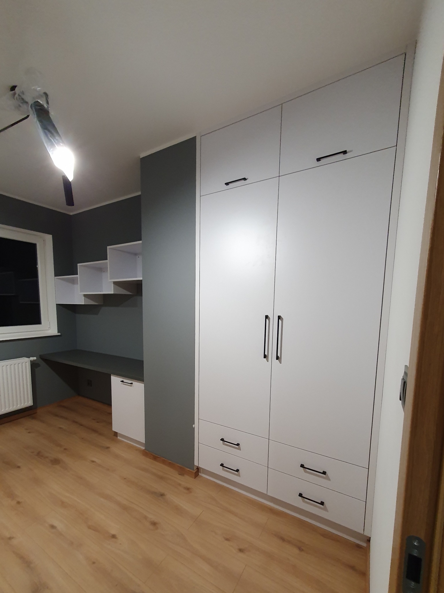 Biała szafa z czarnymi uchwytami, biurko z półkami w pokoju z szarymi ścianami i drewnianą podłogą, widok na sufit z lampą w folii.