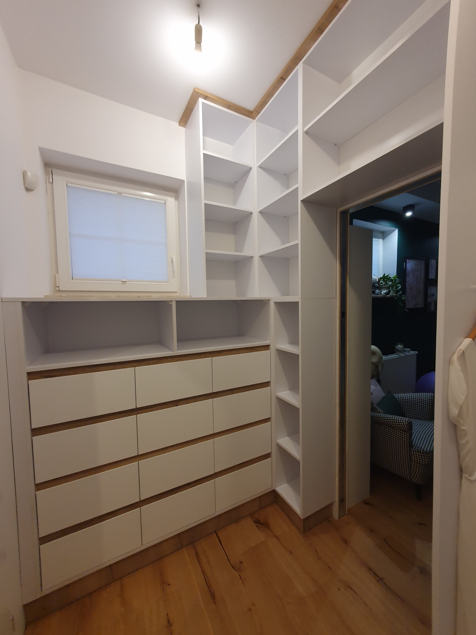 Biała garderoba z szufladami i półkami, drewniane akcenty, widok na wnętrze pokoju z fotelami w tle.