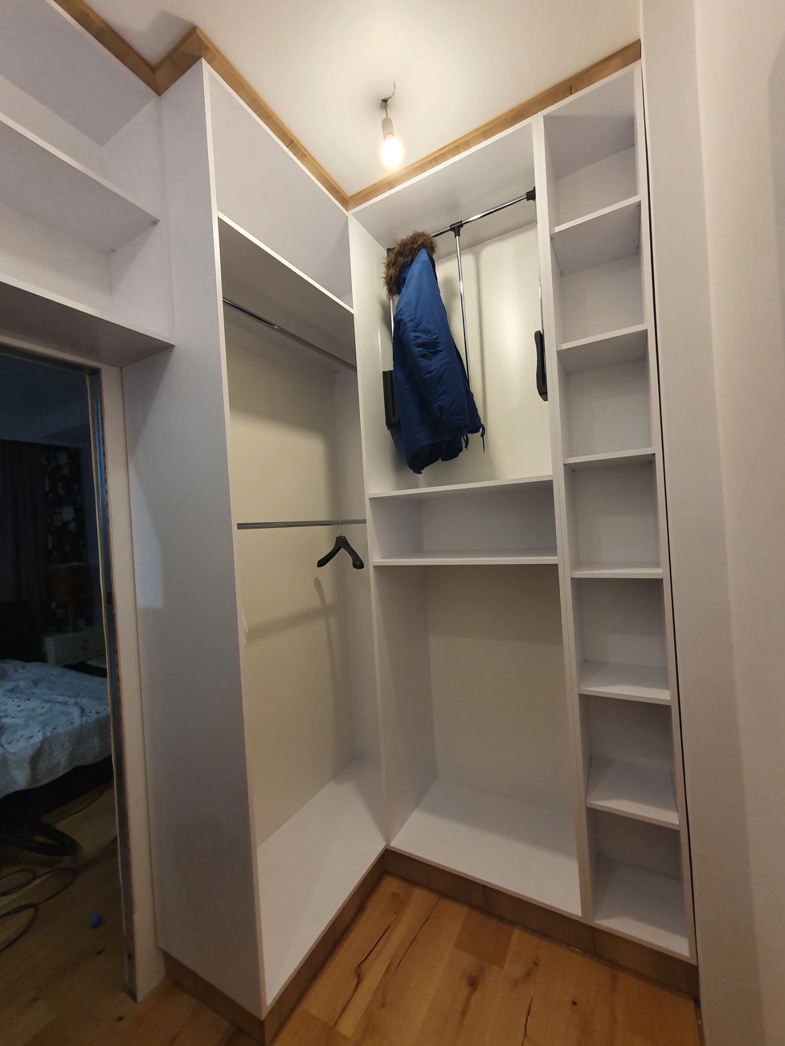 Biała szafa narożna z półkami i drążkami, widoczny fragment pokoju z łóżkiem w tle.