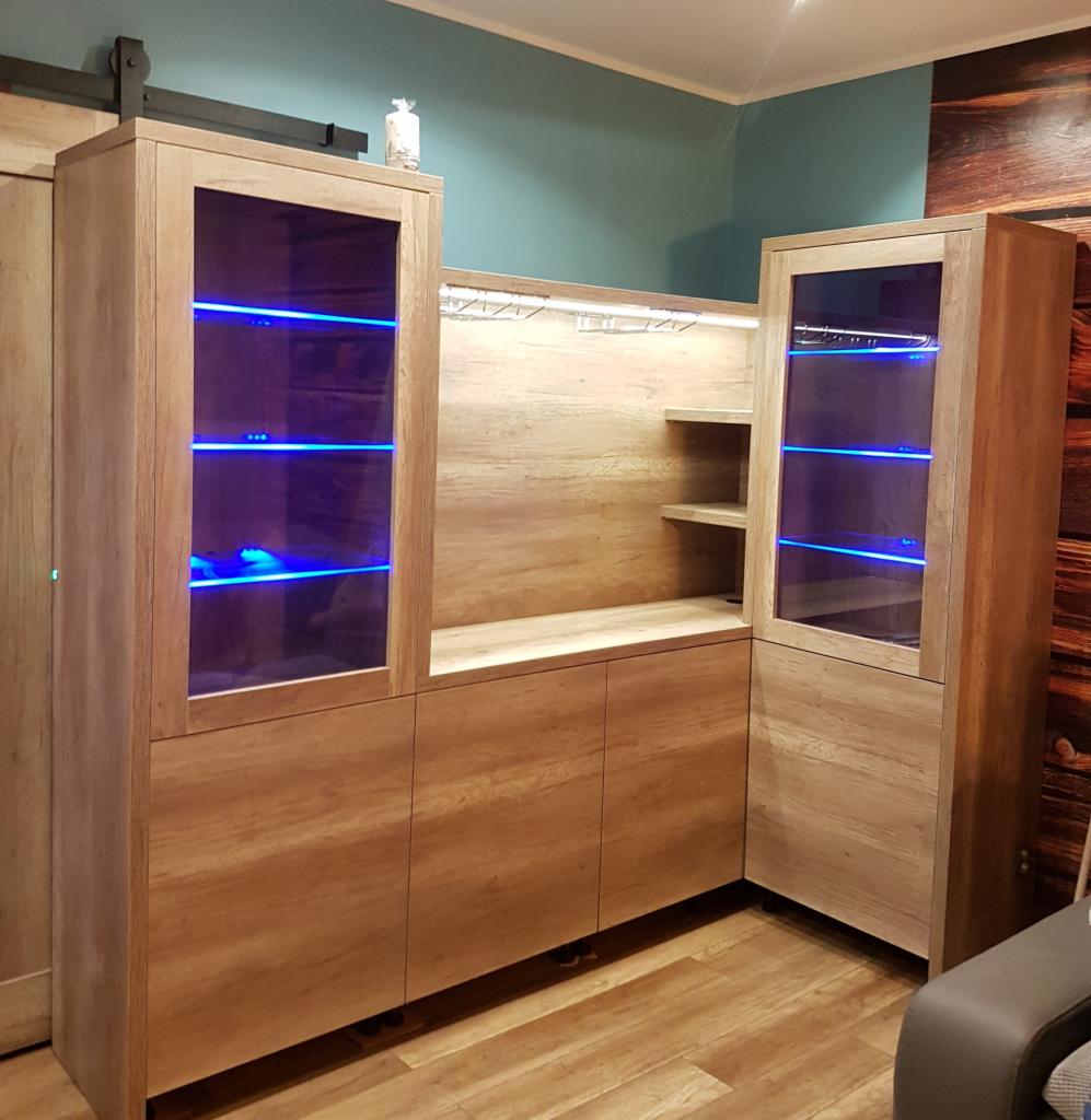 Zabudowa salonu z drewna w odcieniach beżu, z przeszklonymi witrynami podświetlonymi niebieskim LED, półki na kieliszki, widoczne uchwyty na drzwi przesuwne.