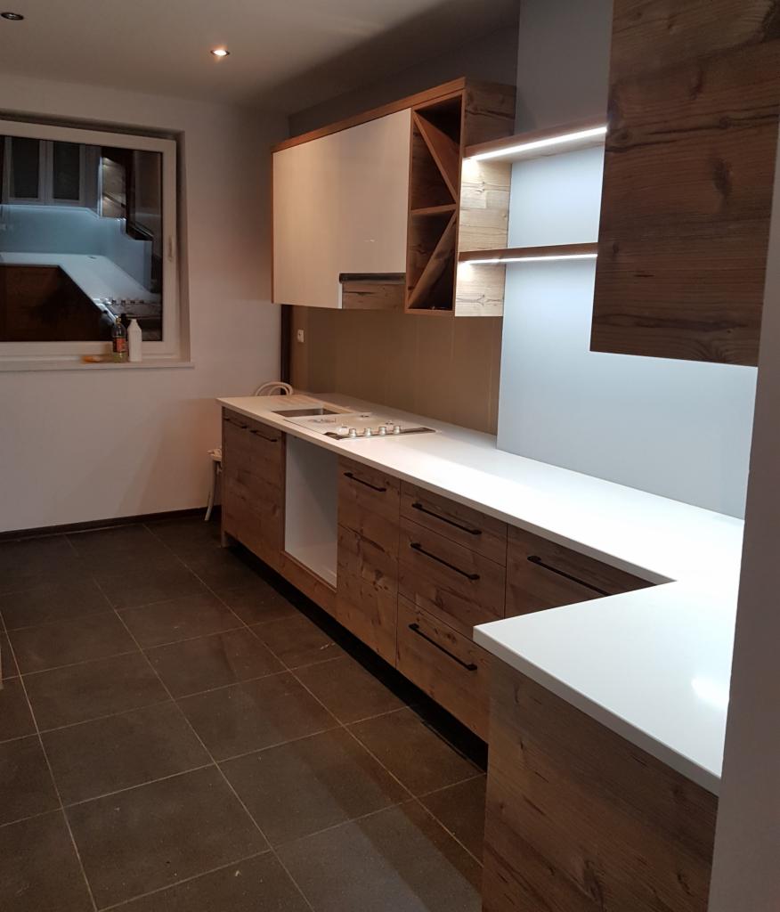 Nowoczesna kuchnia z drewnianymi frontami szafek i białym blatem, oświetlenie LED pod szafkami wiszącymi, widok na okno z widocznym wnętrzem innego pomieszczenia.