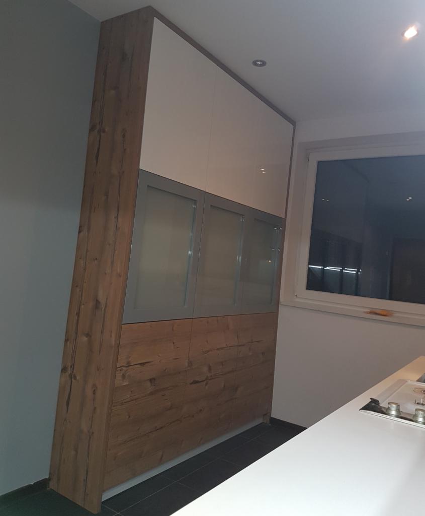 Wysoka szafa wnękowa z drewnianymi bokami, białymi górnymi frontami i szarymi, przeszklonymi drzwiczkami, widoczna w kuchni.