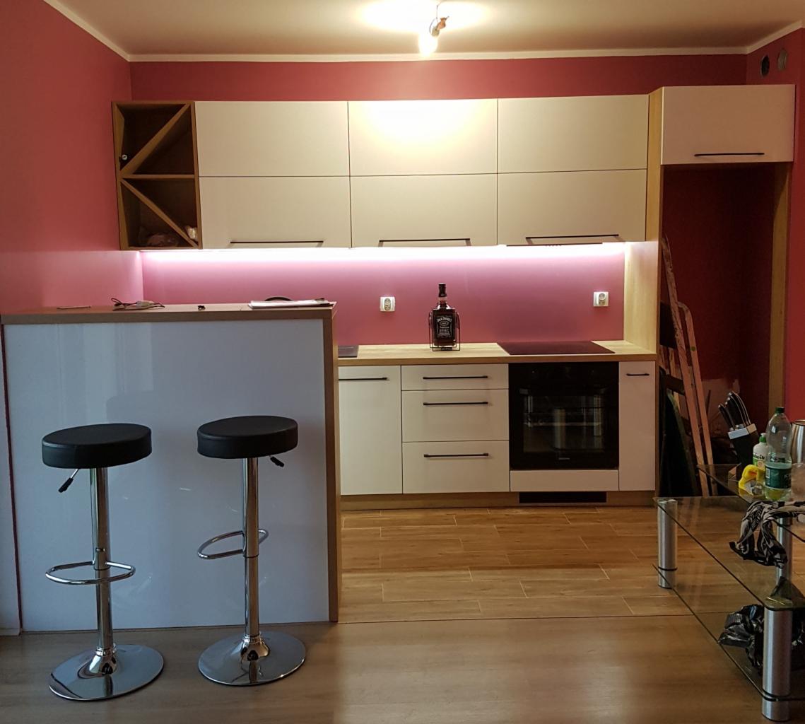 Nowoczesna kuchnia z białymi frontami szafek, podświetleniem LED, blatem drewnianym i barkiem z dwoma hokerami na tle różowych ścian. Na blacie stoi butelka Jack Daniel's.