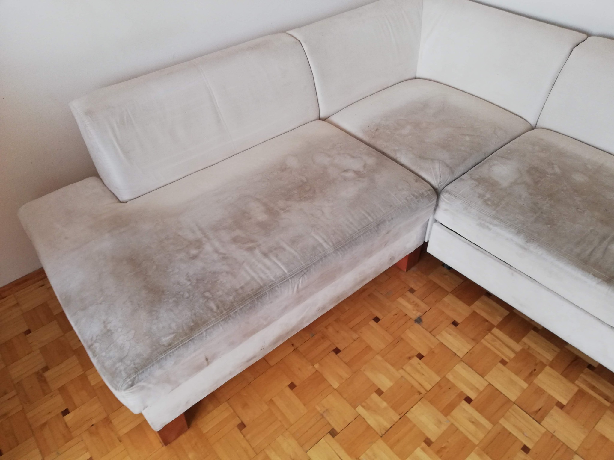 Narożna, jasna kanapa z widocznymi, rozległymi plamami na siedzisku, stojąca na parkiecie ułożonym we wzór jodełki.