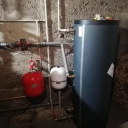 Szary akumulacyjny zbiornik ciepłej wody użytkowej, czerwone naczynie przeponowe i białe naczynie wyrównawcze, zainstalowane w piwnicy z surowymi ścianami, z widocznymi rurami instalacji grzewczej.