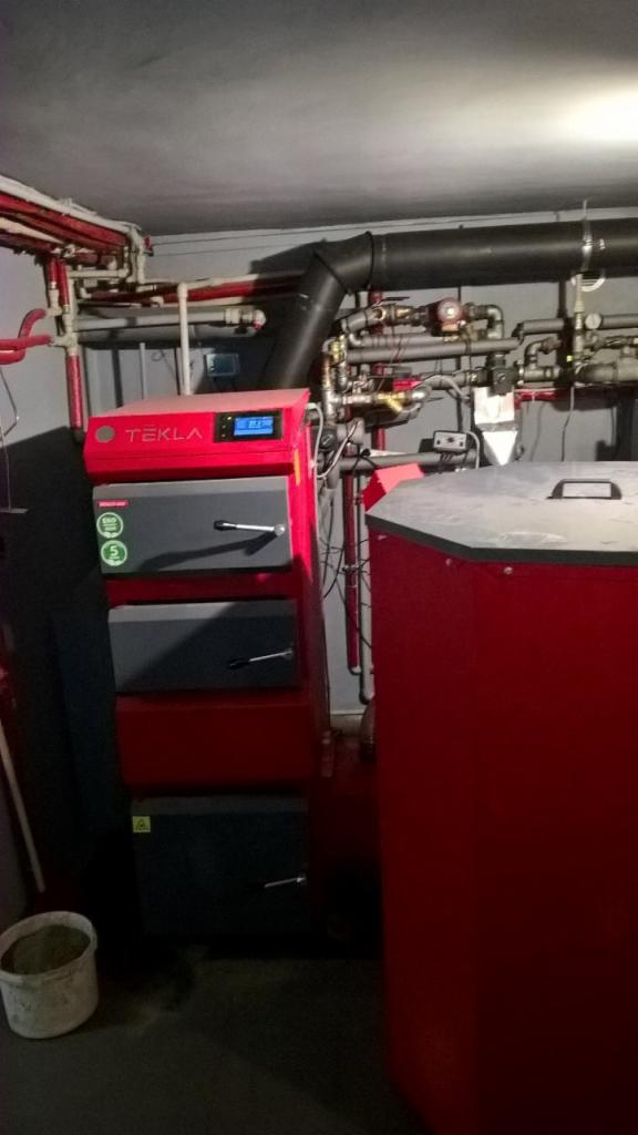 Czerwony piec CO marki Tekla z szarymi drzwiczkami, widoczne rury instalacji grzewczej pod sufitem, instalacja w kotłowni.