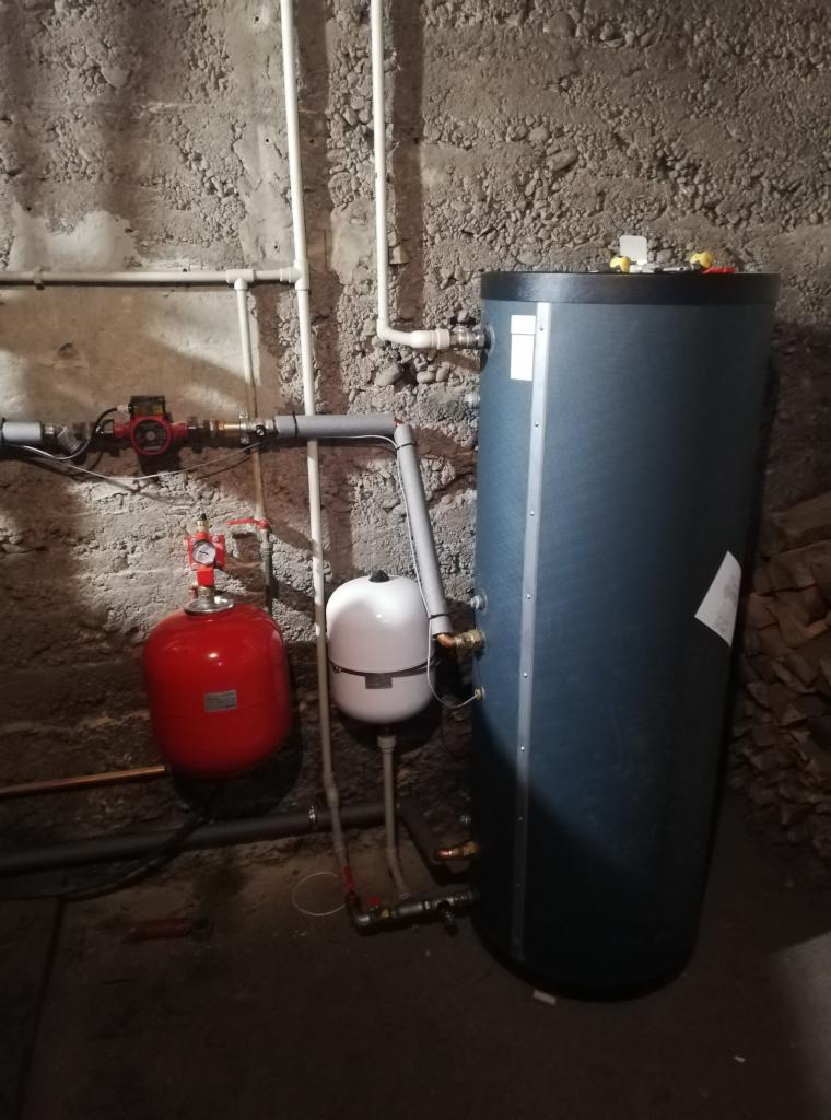 Szary akumulacyjny zbiornik ciepłej wody użytkowej, czerwone naczynie przeponowe i białe naczynie wyrównawcze, zainstalowane w piwnicy z surowymi ścianami, z widocznymi rurami instalacji grzewczej.