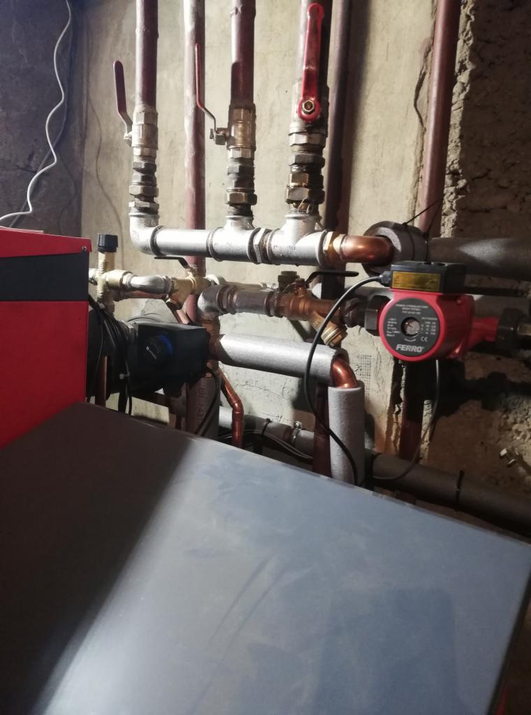 Część instalacji grzewczej z kotłem, rurami miedzianymi i zaworami, widoczna pompa obiegowa Ferro z czerwonym korpusem, zamontowana na tle surowej ściany piwnicy.