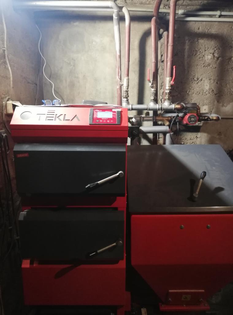 Czerwono-czarny piec Tekla z widocznym panelem sterowania, rurami instalacji grzewczej i zaworami w surowym pomieszczeniu kotłowni.