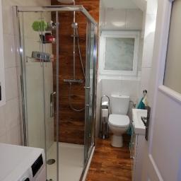 DIMAX SPÓŁKA Z OGRANICZONĄ ODPOWIEDZIALNOŚCIĄ - Wnętrze łazienki po remoncie: kabina prysznicowa z panelami imitującymi drewno, toaleta, umywalka z szafką, pralka z przodu oraz drewnopodobna podłoga.