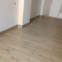 DIMAX SPÓŁKA Z OGRANICZONĄ ODPOWIEDZIALNOŚCIĄ - Ułożona podłoga z jasnych płytek imitujących drewno, widoczne fugi, listwy przypodłogowe i fragment ściany w trakcie remontu.