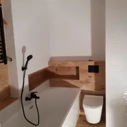 Nowoczesna łazienka z wanną, toaletą i armaturą w kolorze czarnym, wykończona białymi płytkami i panelami imitującymi drewno, oświetlenie sufitowe punktowe.