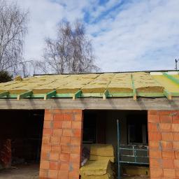 DIMAX SPÓŁKA Z OGRANICZONĄ ODPOWIEDZIALNOŚCIĄ - Budowa domu w trakcie realizacji: ściany z czerwonej cegły, izolacja z wełny mineralnej na dachu, widoczne drewniane krokwie i membrana dachowa, błękitne niebo w tle.