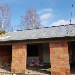 DIMAX SPÓŁKA Z OGRANICZONĄ ODPOWIEDZIALNOŚCIĄ - Budowa domu z czerwonej cegły, widoczny dach w trakcie krycia z membraną i łatami, na tle błękitnego nieba i drzew bez liści.