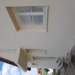 DIMAX SPÓŁKA Z OGRANICZONĄ ODPOWIEDZIALNOŚCIĄ - Świeżo otynkowana elewacja domu jednorodzinnego z zakrytymi folią oknami, widoczne detale wykończenia wokół okien i fragment niezagospodarowanego terenu przy budynku.