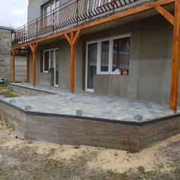 DIMAX SPÓŁKA Z OGRANICZONĄ ODPOWIEDZIALNOŚCIĄ - Szara elewacja surowego domu z drewnianą konstrukcją zadaszenia tarasu i ułożoną kostką brukową na podwyższonym fundamencie, widok z poziomu gruntu.