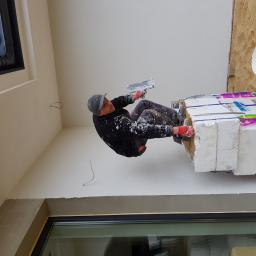 DIMAX SPÓŁKA Z OGRANICZONĄ ODPOWIEDZIALNOŚCIĄ - Pracownik w ubraniu roboczym zacierający gładź na ścianie wewnętrznej, stojąc na bloczkach z betonu komórkowego w nowo wybudowanym domu z oknami dachowymi.