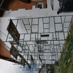 DIMAX SPÓŁKA Z OGRANICZONĄ ODPOWIEDZIALNOŚCIĄ - Dom z cegły z ociepleniem styropianowym w trakcie montażu, widoczne rusztowania, fragment dachu z rynną i okno.