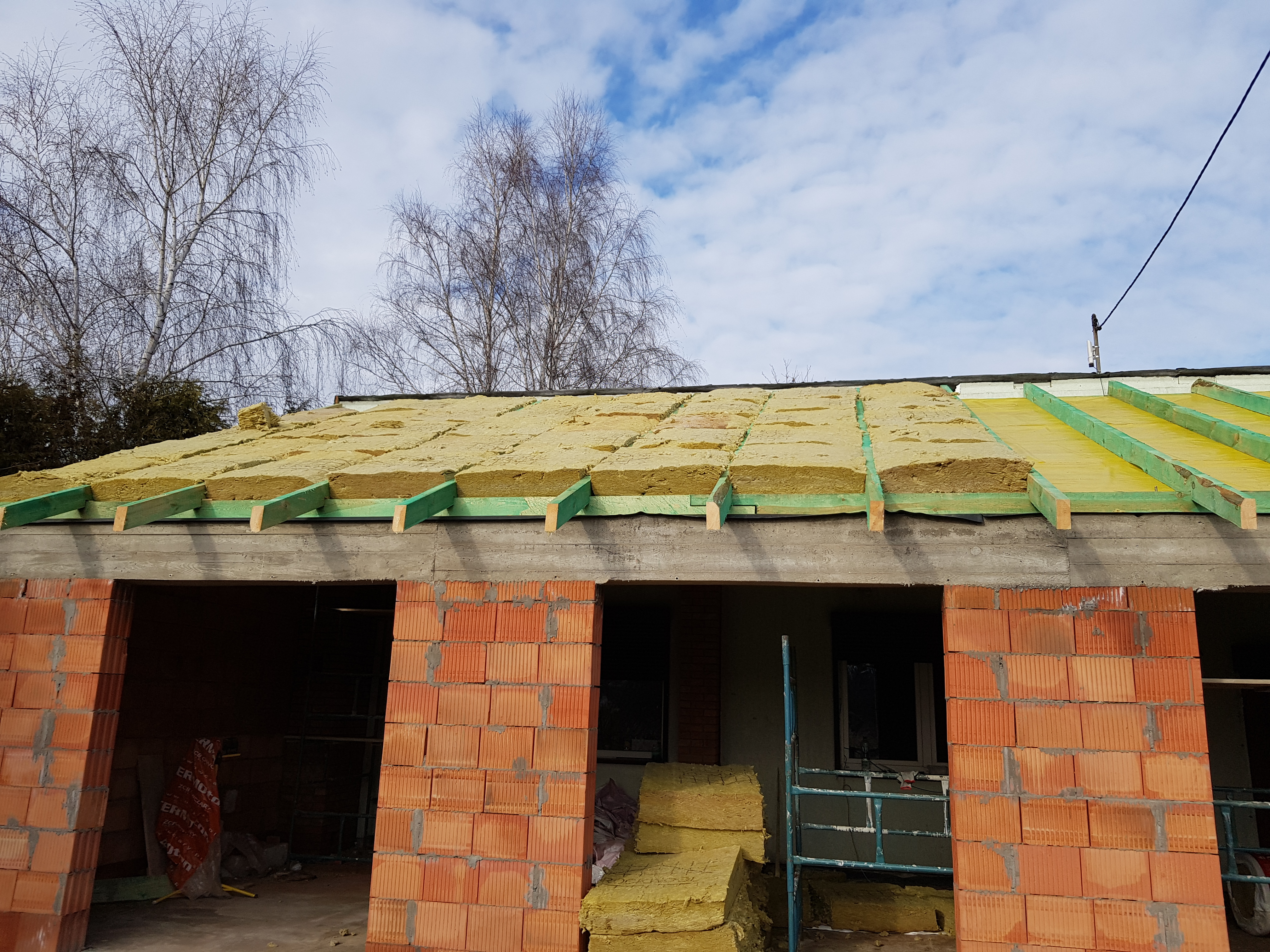 Budowa domu w trakcie realizacji: ściany z czerwonej cegły, izolacja z wełny mineralnej na dachu, widoczne drewniane krokwie i membrana dachowa, błękitne niebo w tle.