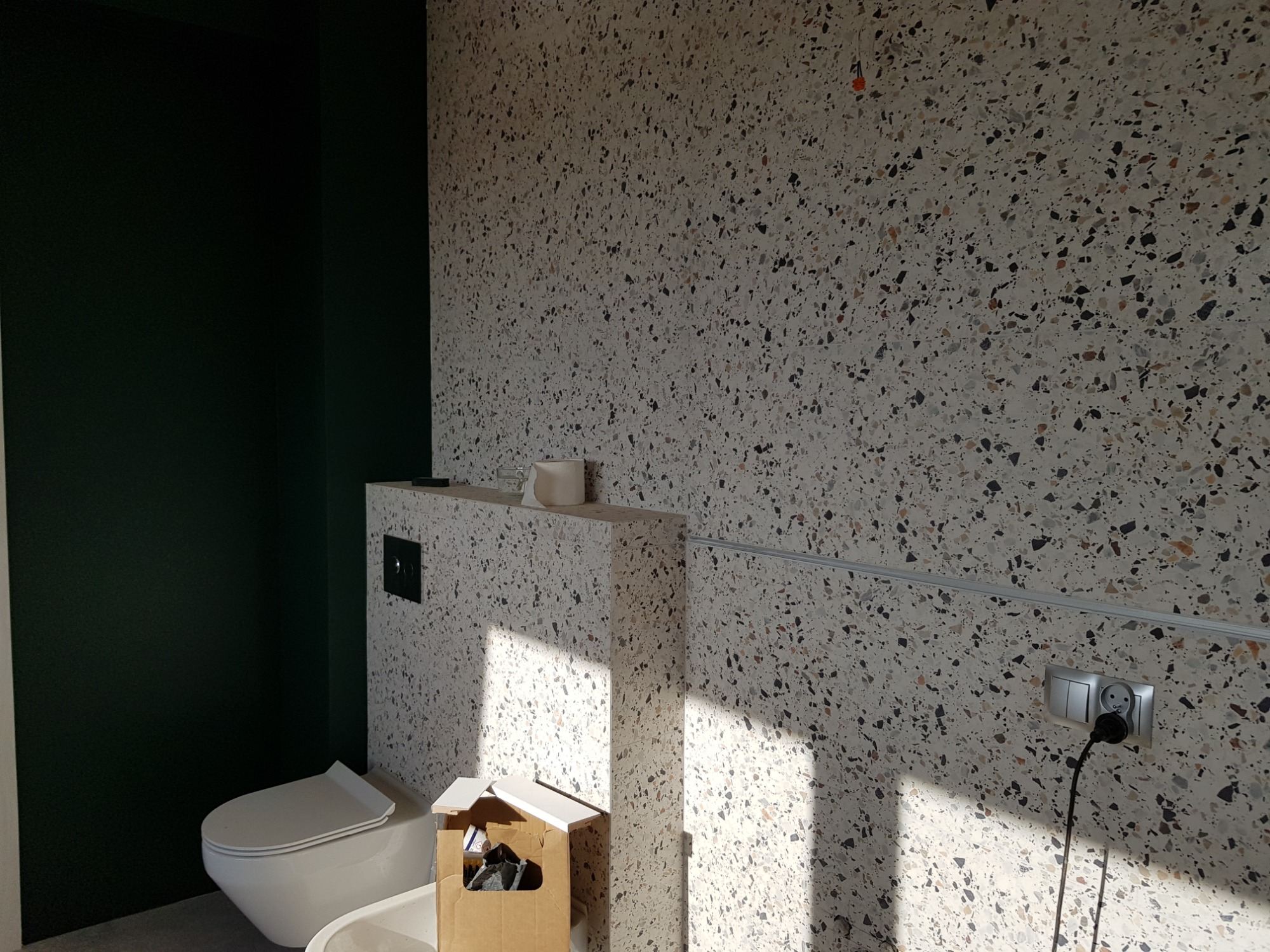 Wnętrze łazienki w trakcie wykańczania, ściany wyłożone płytkami imitującymi lastryko, widoczny biały sedes, puszka instalacyjna i gniazdko elektryczne z podłączonym kablem, kubek na półce i karton...