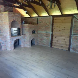 Zadaszona przestrzeń z ceglanym grillem, piecem chlebowym i podłogą z płytek imitujących drewno, oświetlona żarówkami dekoracyjnymi i żyrandolem.