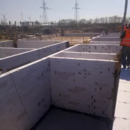 Wykop pod fundamenty z wylanym betonem i wymurowanymi ścianami z białego bloczka, widoczne zbrojenia, w tle słupy wysokiego napięcia.