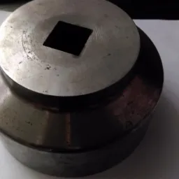 Metalowy element tokarski z kwadratowym otworem, widoczne ślady użytkowania i drobne zarysowania na powierzchni.