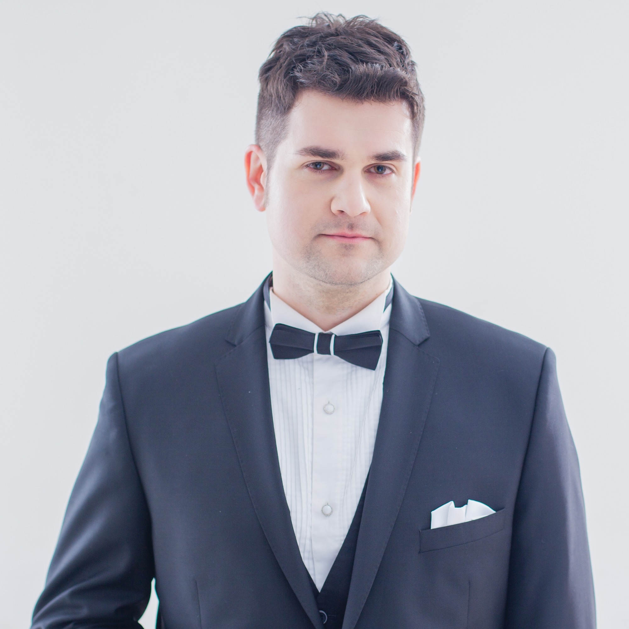 Portret mężczyzny w czarnym smokingu z białą poszetką, na jasnym tle.