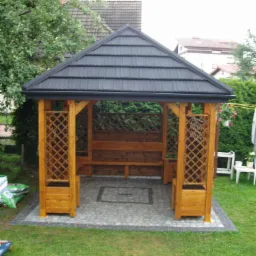 Drewniana altana ogrodowa z czarnym dachem, wykończona kostką brukową i kratownicą. Widok na zadbany trawnik i zieleń. Elementy architektury ogrodowej.