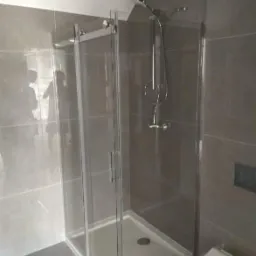 Kabina prysznicowa z przesuwnymi drzwiami, zamontowana w szarej łazience z płytkami, widoczny zestaw natryskowy i fragment toalety.