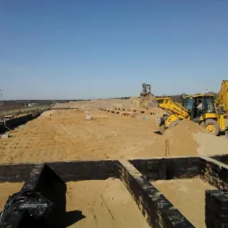 Wykop pod fundamenty domu jednorodzinnego z widoczną izolacją pionową, w tle pracująca koparko-ładowarka Komatsu i wywrotka, teren wyrównany, przygotowany do dalszych prac.