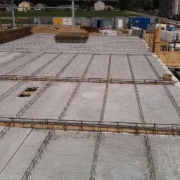 Betonowy strop Teriva z widocznym zbrojeniem i otworem, na tle plac budowy z kontenerami i dźwigiem.