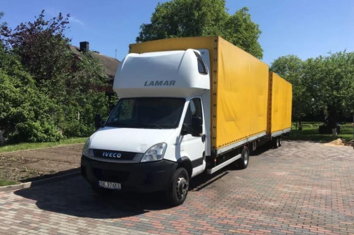 Biała ciężarówka Iveco z żółtą plandeką i napisem 'LAMAR' na kabinie, zaparkowana na brukowanej nawierzchni w słoneczny dzień.